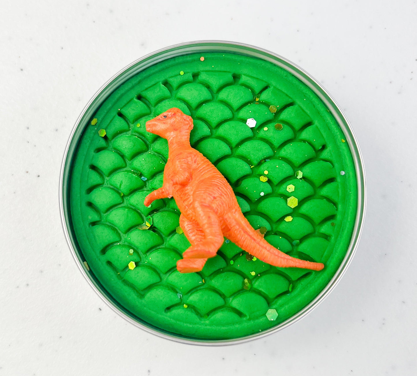 Green dinosaur skin print playdough pot with a small orange dinosaur toy on a white background