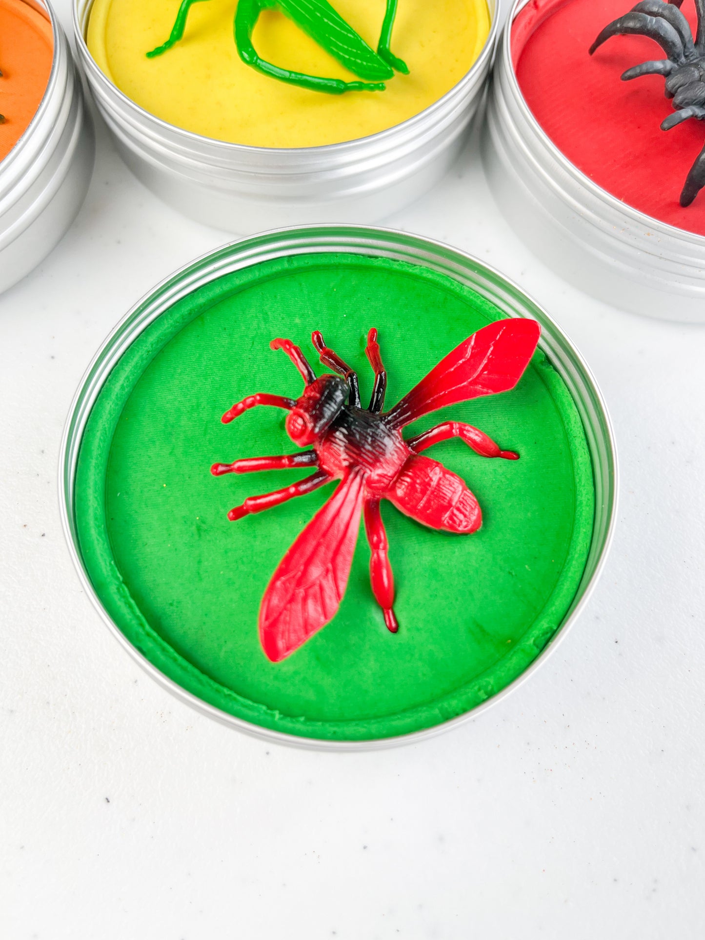 Insect Themed Sensory Playdough Pots for Alternative Party Bag Fillers. Eco friendly, handmade playdough in a mix of colours with reptile animal characters on top. One tin of green playdough with fly figure on top