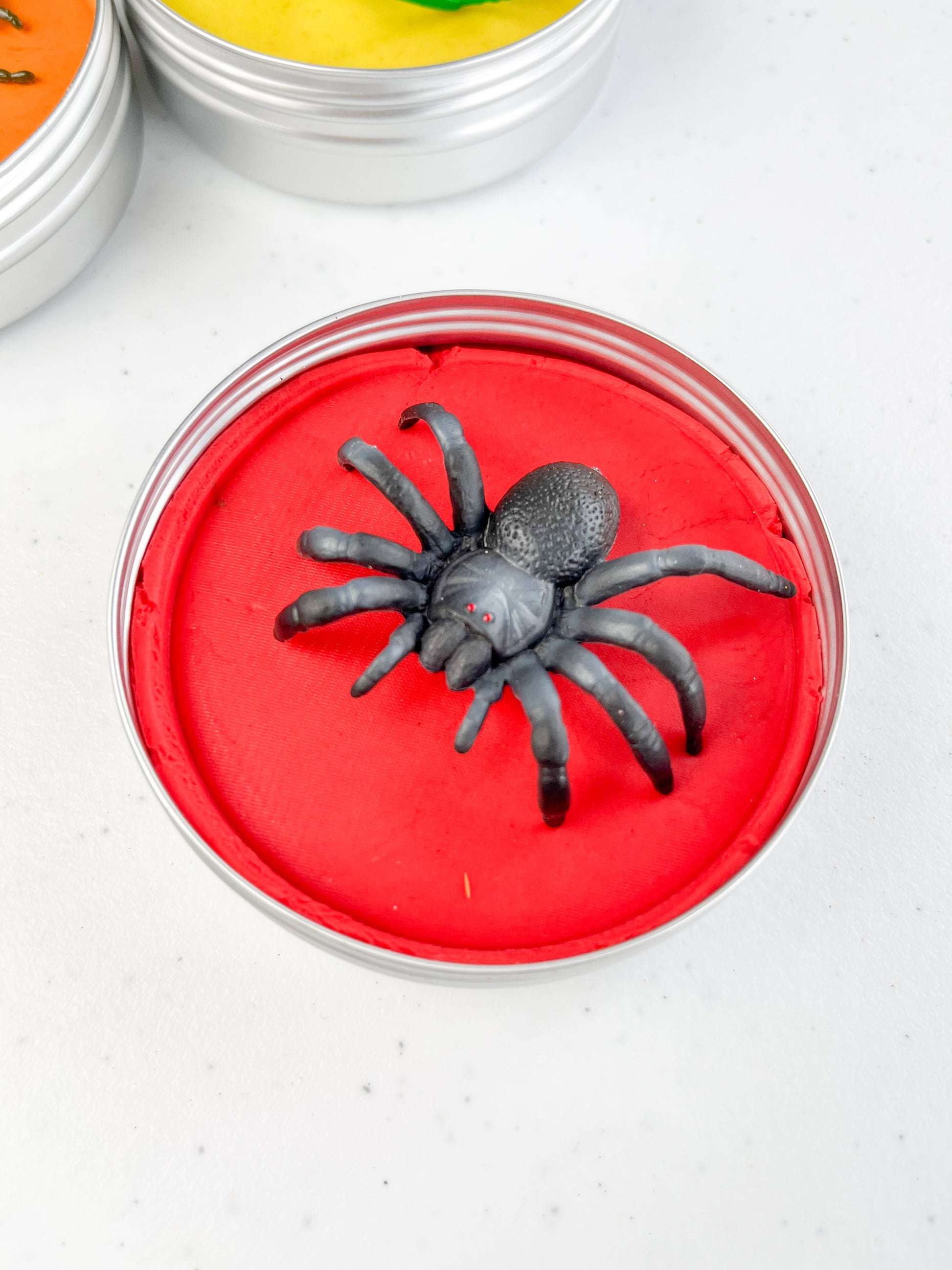 Insect Themed Sensory Playdough Pots for Alternative Party Bag Fillers. Eco friendly, handmade playdough in a mix of colours with reptile animal characters on top. One tin of red playdough with spider figure on top