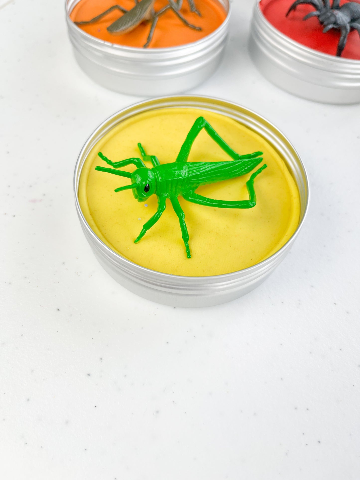 Insect Themed Sensory Playdough Pots for Alternative Party Bag Fillers. Eco friendly, handmade playdough in a mix of colours with reptile animal characters on top. One tin of yellow playdough with grasshopper figure on top