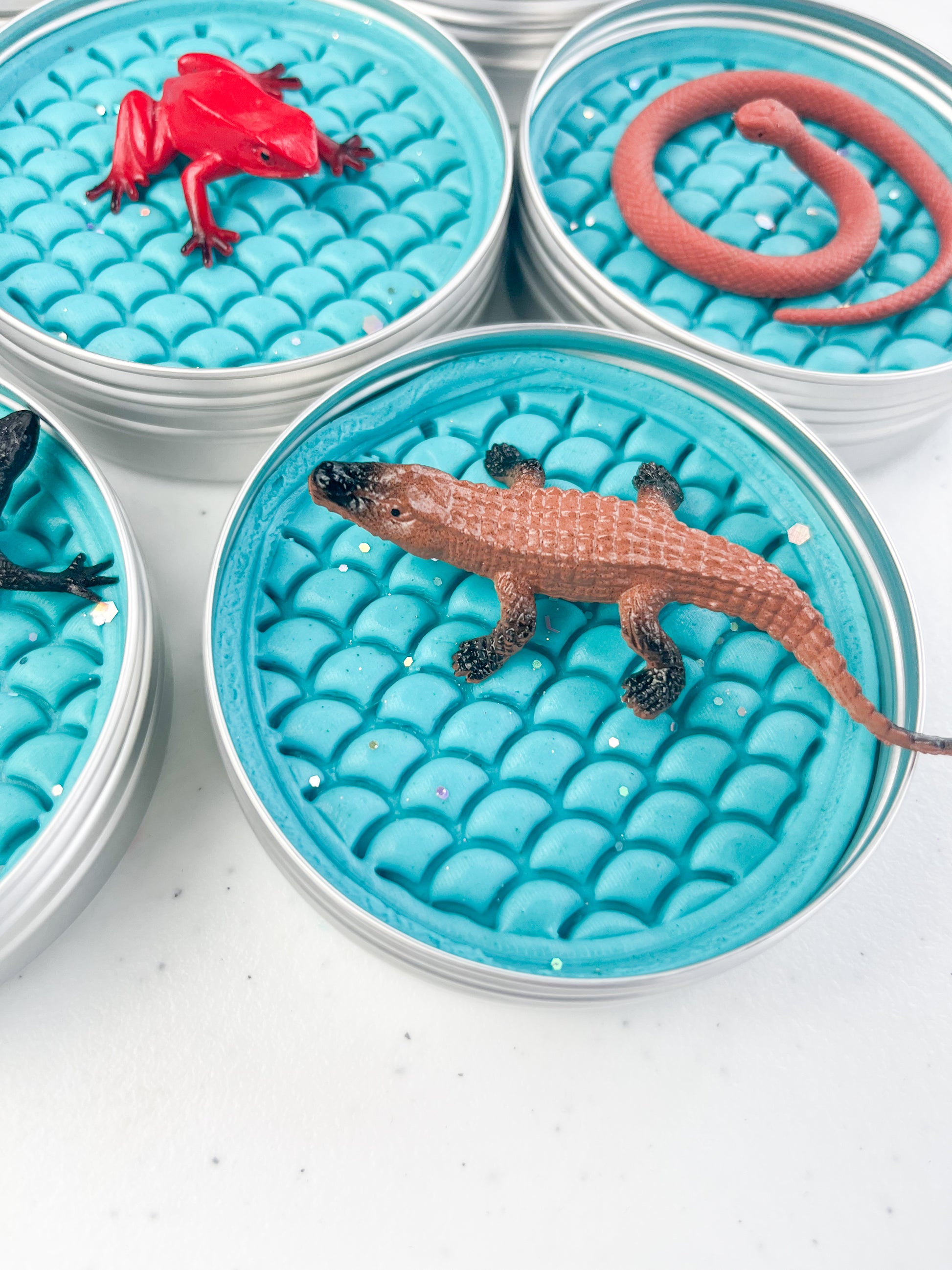 Reptile Themed Sensory Playdough Pots for Alternative Party Bag Fillers. Eco friendly, handmade playdough in a mix of colours with reptile animal characters on top. Set of blue playdough pots with various reptile figures on top
