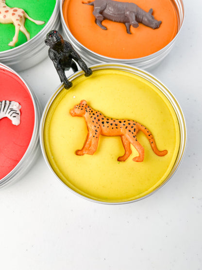 Safari Animal Themed Sensory Playdough Pots for Alternative Party Bag Fillers. Eco friendly, handmade playdough in a mix of colours with safari animal characters on top. Set of primary coloured playdough pots with various animal figures on top. Yellow Playdough in metal tub with cheetah figure on top.