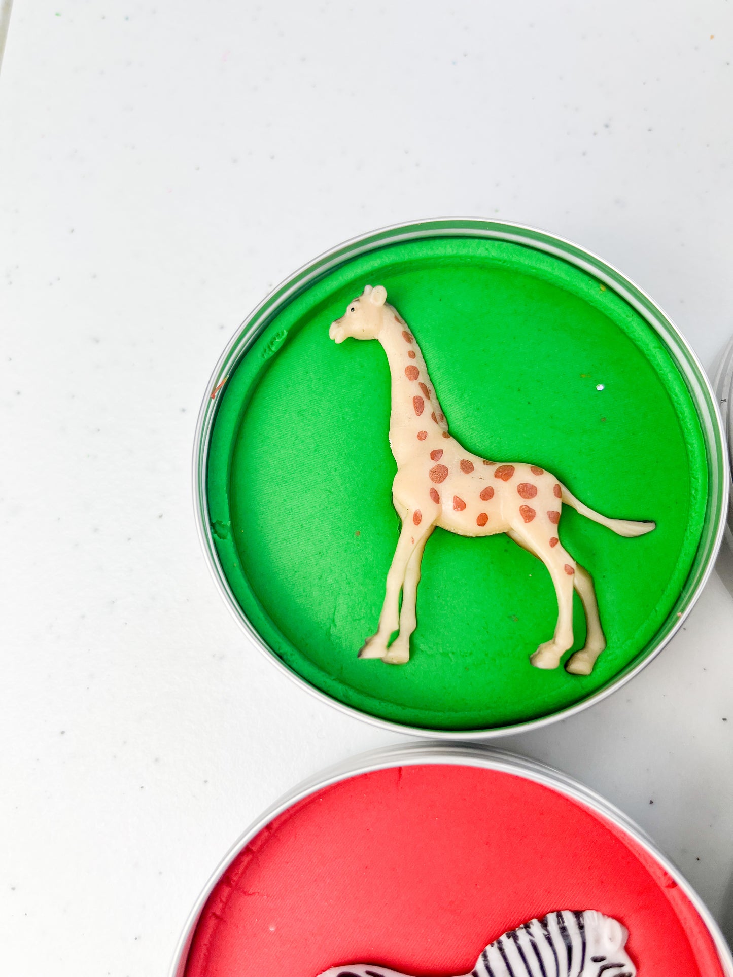 Safari Animal Themed Sensory Playdough Pots for Alternative Party Bag Fillers. Eco friendly, handmade playdough in a mix of colours with safari animal characters on top. Set of primary coloured playdough pots with various animal figures on top. Green Playdough in metal tub with giraffe figure on top.