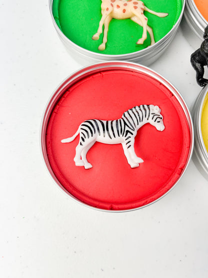 Safari Animal Themed Sensory Playdough Pots for Alternative Party Bag Fillers. Eco friendly, handmade playdough in a mix of colours with safari animal characters on top. Set of primary coloured playdough pots with various animal figures on top. Red Playdough in metal tub with zebra figure on top.