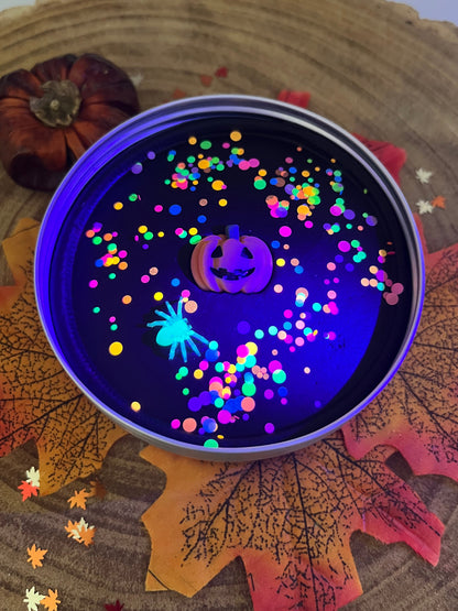 A round tin filled with glowing multicolor confetti and a neon spider on a dark background, styled on autumn leaves and a wood slice with a small pumpkin prop.