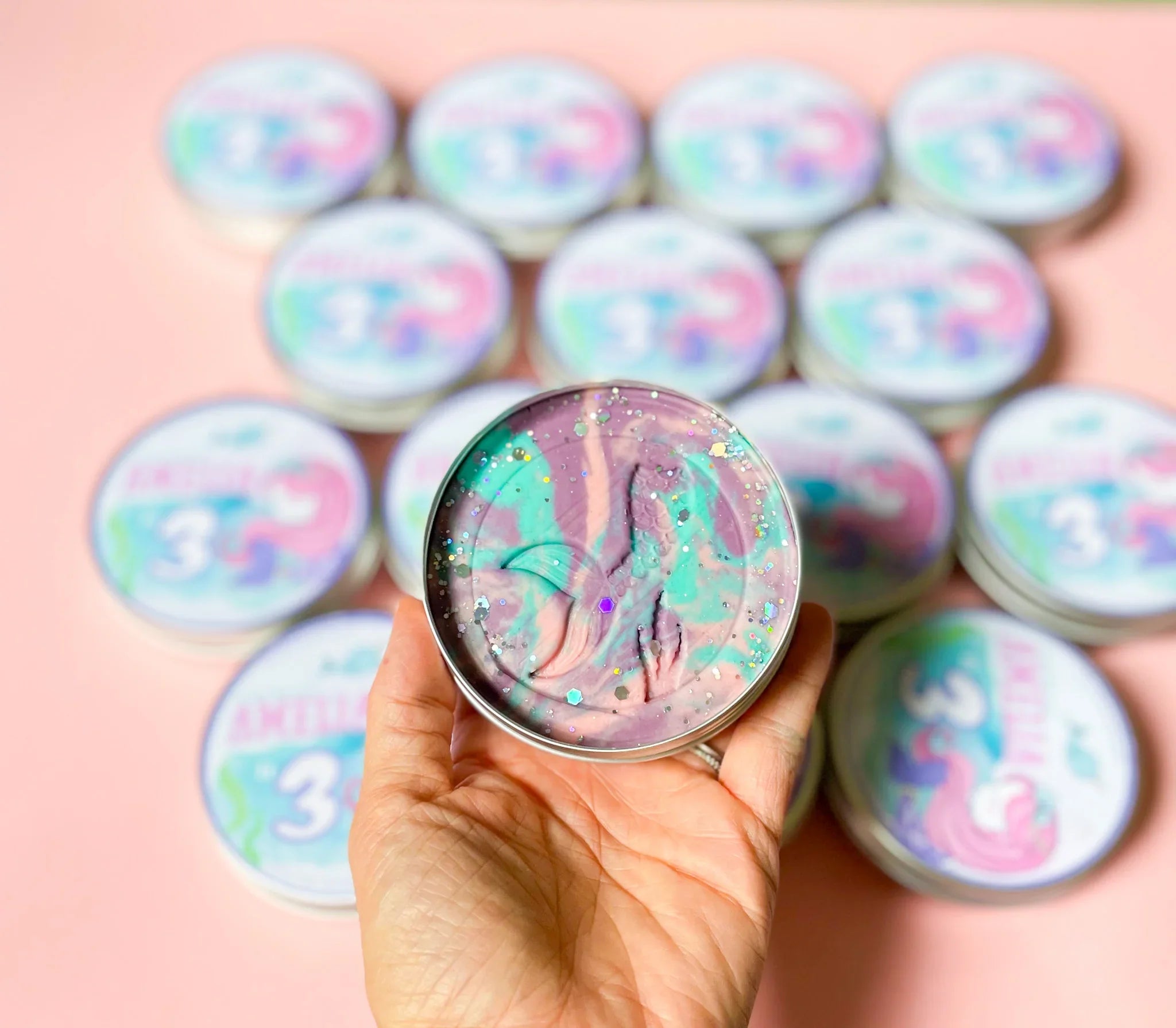 A hand holding a round tin of mermaid-themed playdough with a colorful, sparkly design on a pink background, with the number 3 visible on the tin for personalised eco friendly party bag fillers